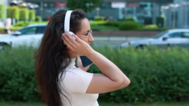 Neşeli, olumlu, beyaz kulaklıkla müzik dinleyen, elinde akıllı telefonuyla şehirde şarkı söyleyen güzel bir kadının yan görüntüsü. Kapatın. Sokakta şarkı söyleyen neşeli genç bir kadın.