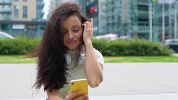 与快乐的白种人漂亮的女性亲密接触 耳机坐在城市里 在智能手机上用社交媒体愉快地微笑 年轻快乐的女人用小玩意 休闲活动城市生活 — 图库视频影像