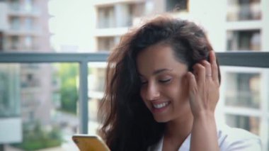 Beyaz bornozlu beyaz bir kadın balkonda oturuyor ve iyi bir ruh hali içinde cep telefonuyla internette geziniyor. Akıllı telefona bakarken gülümseyen güzel, neşeli bir bayan. Boş zaman konsepti