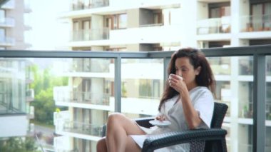 Beyaz bir kadın apartmanın balkonunda oturmuş kahvenin tadını çıkarıyor. Evdeki balkonda dinlenen koyu renk saçlı güzel bir kadın. Boş zaman.