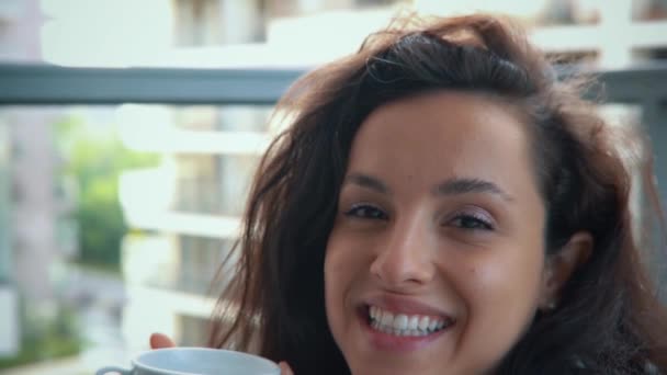 穿着白袍的美丽迷人的年轻女子坐在阳台上 喝着一杯热饮 白种人黑发快乐的家庭主妇在公寓的阳台上喝咖啡 对着相机微笑 — 图库视频影像