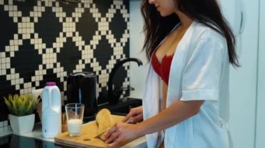 Kırmızı iç çamaşırlı güzel bir kadın mutfakta duruyor ve kahvaltı hazırlayan bir baget kesiyor. Ağır çekim. Koyu renk saçlı, seksi iç çamaşırlı kadın ev kadınına yakın çekim yemek hazırlar.