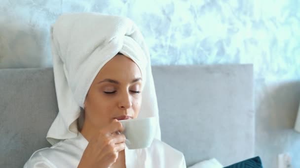 穿着浴袍 头戴毛巾的漂亮快乐女人在洗澡后躺在床上喝咖啡 享受放松的时光 美丽的白人女性躺在舒适的家的卧室里 慢动作 — 图库视频影像
