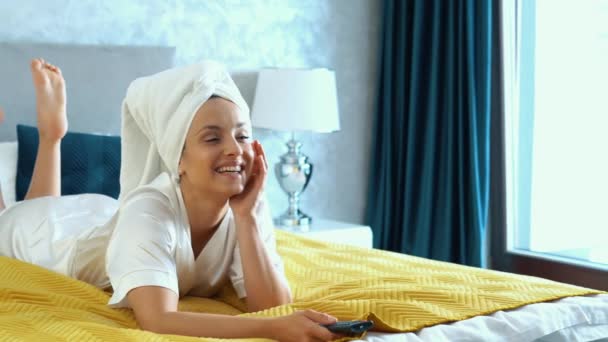 テレビを見て微笑んだ後 自宅のベッドの室内にタオルを頭に横たわっ白人の美しい若い女性 スローモーション リモートコントロールを使用して幸せな女性の時計テレビのチャンネルを変更 — ストック動画