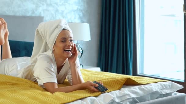 白种人快乐的年轻女人 洗完澡后 头戴毛巾躺在床上看电视上的喜剧电影 休闲活动快乐的女性看电视 笑着拿着遥控装置 慢动作 — 图库视频影像