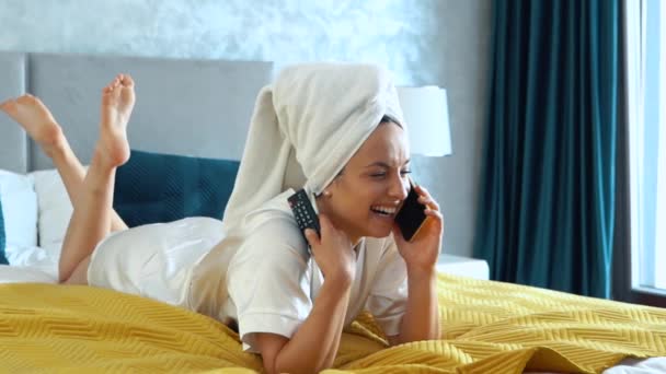 快乐的年轻女人 头戴毛巾 面带微笑 在智能手机上说话 洗完澡后躺在床上看电视 休闲的概念 快乐的女士看电视和打电话 靠近点 — 图库视频影像
