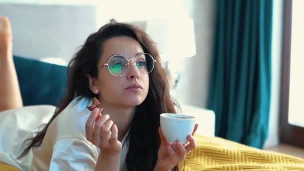 テレビを見ている白いローブの若い女性が怠惰な朝を過ごし コーヒーを飲みながらマカロンを食べています クッキーでコーヒーを飲むメガネのブルネットの魅力的な女性のクローズアップ — ストック動画