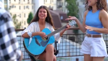 Gençler bir partide eğlenirken çatıda gitar çalan güzel bir genç kadın. Neşeli, pozitif bayan müzisyen enstrümanda çalıyor. Çatı partisinde dans eden gençler. Yaz eğlencesi