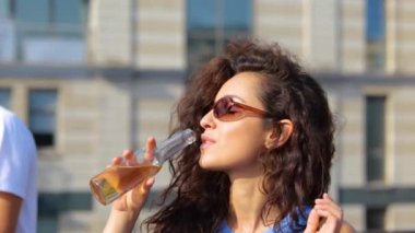 Güneşli bir günde açık havada bira içen, güneş gözlüklü, kıvırcık saçlı, güzel beyaz bir kadın. İçki dansı yapan mutlu, şık bir kadın. İnsanlar arka planda dans eder