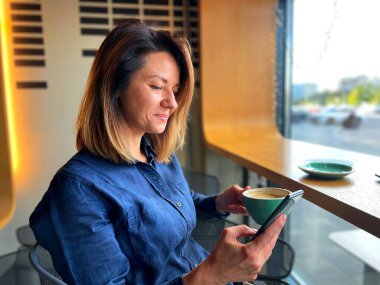 Kafeteryada bir fincan kahveyle oturan ve internette akıllı telefon kullanan genç bayanı yakından çek. Güzel beyaz kadın kahve içer ve cep telefonu ekranına bakar. Boş zaman konsepti