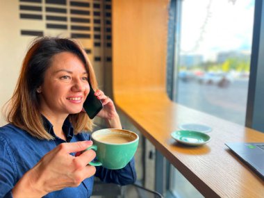 Restoranda pencerenin önünde oturup kahve içerken cep telefonuyla konuşan mutlu güzel bir kadın. Boş zaman. Kadın cep telefonuyla arıyor. Konuşma konsepti