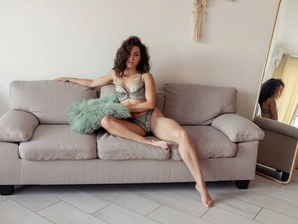 Modern odadaki bir kanepede oturmuş kameraya bakan seksi iç çamaşırlı güzel beyaz kadın. Çekici, siyah saçlı bir kadın. Kanepede, iç çamaşırlarıyla yastık tutuyor.