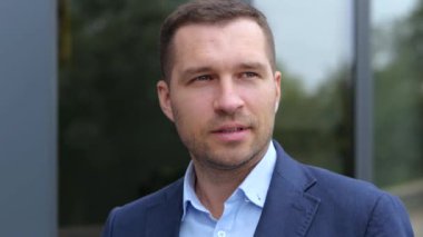 Takım elbiseli, yakışıklı ve kendine güvenen genç bir adam şehirde dolanıp etrafa bakıyor. Kafkasyalı başarılı iş adamı sokakta yürüyor. Şehir tarzı. Ağır çekim. İş konsepti.