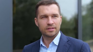 Takım elbiseli, yakışıklı ve kendine güvenen genç bir adam şehirde dolanıp etrafa bakıyor. Kafkasyalı başarılı iş adamı sokakta yürüyor. Şehir tarzı. Ağır çekim. İş konsepti.