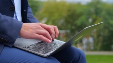 Genç adamın dizüstü bilgisayarda çalıştığı ve klavyeyle parkta oturduğu görüntüleri kapatın. Sokakta bilgisayarda yazan erkek parmakları. İş hayatı. İşçi konsepti. Yavaş çekim