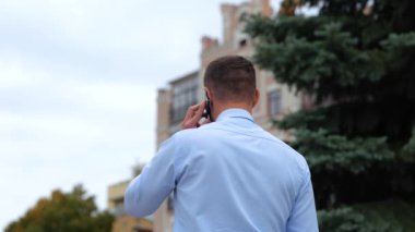 Sokakta yürüyen ve cep telefonuyla konuşan gömlekli bir adamın arka görüntüsü. Şehirdeki bir adam tek başına yürüyor ve cep telefonuyla konuşuyor. İletişim. Telefon görüşmesi konsepti. Ağır çekim. Kapat.