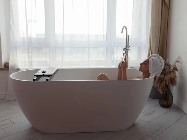 Side view of relaxed young Caucasian woman lying in modern bathtub with towel on head texting on smartphone. Sexy female browsing online on mobile phone while taking bath at home. Relax and leisure