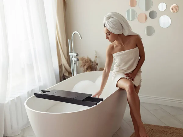 Beautiful Caucasian woman in towel sitting on bathtub with bubbles preparing a bath. Pretty young female in modern bathroom touching water in tub indoor. Woman prepares relaxation bath. Home concept