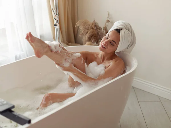 若いかなり陽気な女性は自宅で白い浴槽に石鹸泡で彼女の体を洗う 白人の美しい幸せな女性は 近代的なバスルームで泡風呂に入る 週末は遅い リラックスしたコンセプト — ストック写真