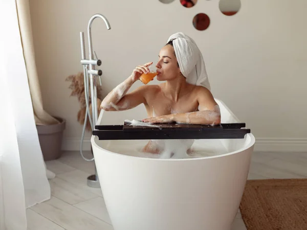 浴槽に座ってオレンジジュースを飲んでいるソープ泡の中に裸のセクシーな可愛い女性 現代の浴槽で白人の美しい女性の入浴 リラクゼーション 豪華なライフスタイル ホテルでの週末 — ストック写真