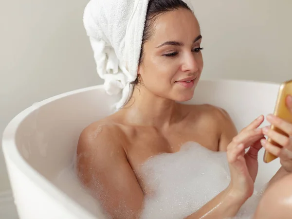 泡と泡がスマートフォンにテキストメッセージで熱い風呂でリラックスした1人の美しい若い女性 笑顔の女性が入浴し 携帯電話サーフィンのインターネットを使用して閉じます — ストック写真
