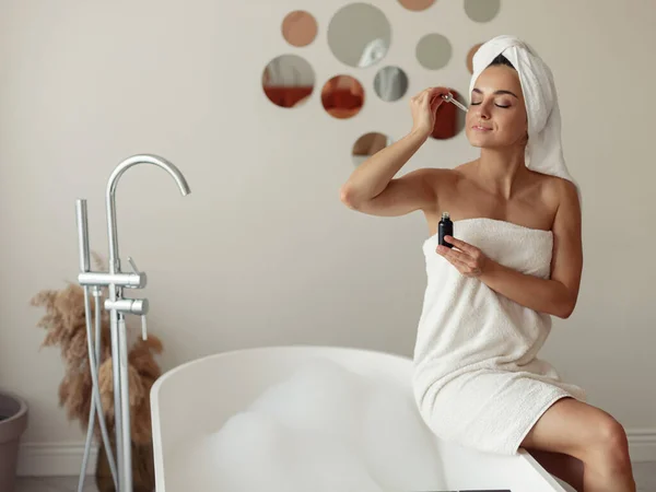 Pretty woman applying face serum using pipette for moisturizing skin in bathroom. Morning skincare routine. Young beautiful female naked in towel sitting on bathtub using beauty product