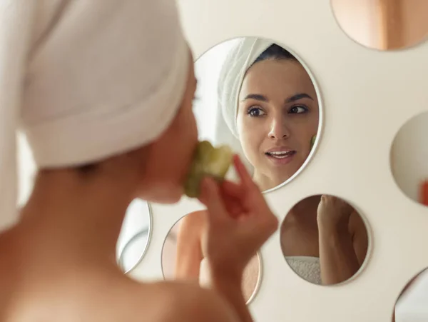 Close up of happy young Caucasian woman doing massage with quartz stone face tool in bathroom looking at mirror. Gua sha acupressure. Skin treatment at home. Anti-aging, skin care concept