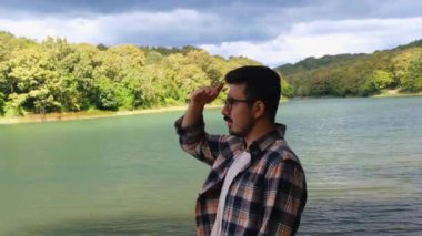 Genç adam elini kullanarak başka tarafa bakıyor, gözlüklü adam göl, orman ve doğa manzarasına bakıyor, 4k 60 fps video, doğadaki konsept görüntüler, ileriye dönük fikir.
