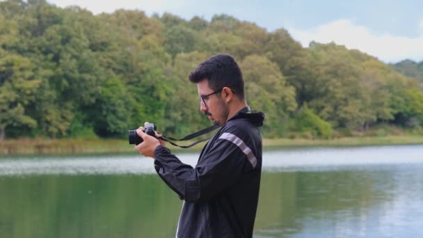 プロのカメラで湖を撮影する若者 ビデオコンセプトを取ります Fps 自然の中でカメラを使用して眼鏡男 曇りの空と美しい風景 森の中での活動とレジャー — ストック動画