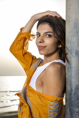 Çekici bir genç esmer, deniz manzarasının önündeki bir direğe yaslanmış çekici bir bakışla başını kameraya çeviriyor. Yaz mevsimi kavramı.