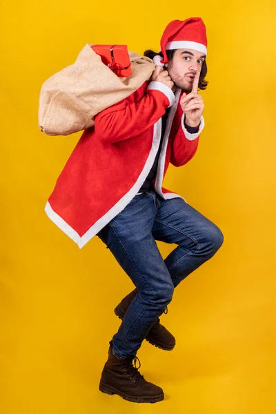 A young handsome man dressed up like Santa Claus with a sack full of presents invites to be silent and keep a secret by placing the index on his lips.