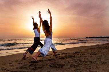 İki genç kadın gün batımında plajda yoga pozu veriyor. Yaz sonu atmosferinde enerji ve dostluk hissi 