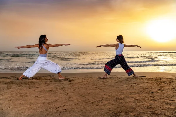 Dos Mujeres Jóvenes Haciendo Yoga Guerrero Posan Playa Atardecer Sentido — Foto de Stock