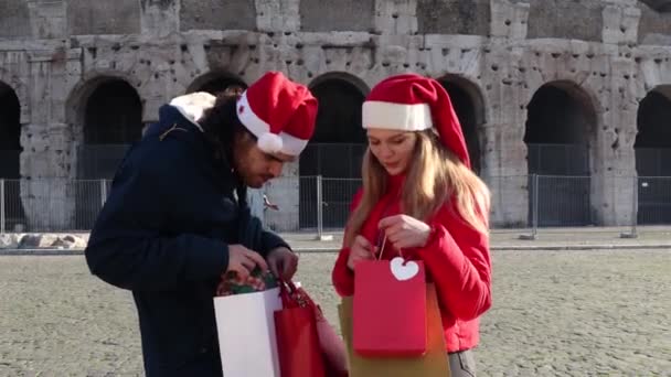 圣诞节购物时间 年轻的夫妇戴着圣诞老人的帽子 站在竞技场前面 正在检查他们买的礼物 — 图库视频影像