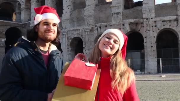 圣诞节购物时间 快乐的年轻夫妇戴着圣诞老人的帽子站在竞技场前 他们笑着展示他们为圣诞节买的礼物 — 图库视频影像