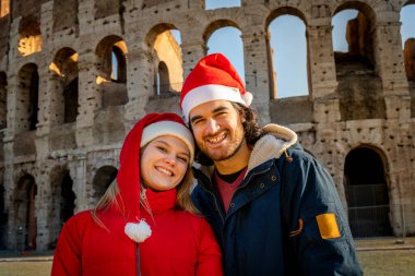 Colosseum 'un önünde kırmızı Noel Baba şapkalarıyla fotoğraf çeken genç bir çift. Mutluluk ve sevgi hissi. Noel tatili konsepti.