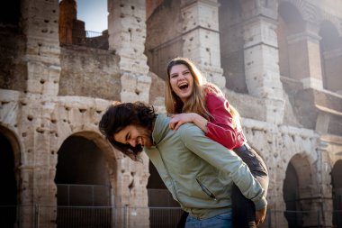 Genç çift Roma 'da seyahat ederken eğleniyor. Adam kız arkadaşını Colosseum 'un önünde sırtında taşıyor..
