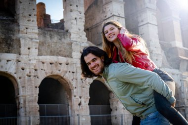 Genç çift Roma 'da seyahat ederken eğleniyor. Adam kız arkadaşını Colosseum 'un önünde sırtında taşıyor..