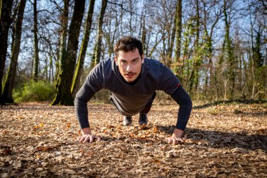 Koşucu, ormanda şınav çekiyor. Mavi gözlü ve bıyıklı genç adam müziği dinlerken yerde şınav çekiyor..
