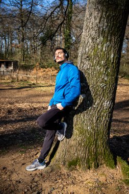 Egzersiz yaparken müzik dinleyen mavi gözlü ve bıyıklı yakışıklı bir gencin portresi. Mavi ceketli adam ormanda antrenman yapıyor ve bir meşe ağacına yaslanıyor..