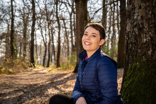 Genç, gülümseyen bir kadın ormandaki bir meşe ağacının dibinde oturuyor. Güzel bir kadın parkta yürüdükten sonra dinleniyor.