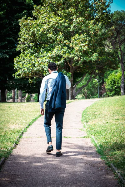 The businessman is walking in the park. The young businessman strolls holding his jacket over his shoulder.