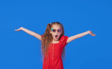 Kalp şeklinde güneş gözlüklü, şaşırmış, küçük kız çocuğunun portresi. Ağzı açık bir çocuk mavi arka planda eğleniyor. Kameraya bakıyorum. Komik surat.