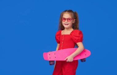 Parlak elbiseli havalı küçük kız kaykayın üzerinde oturur ve mavi arka planda izole edilmiş kameraya bakar.