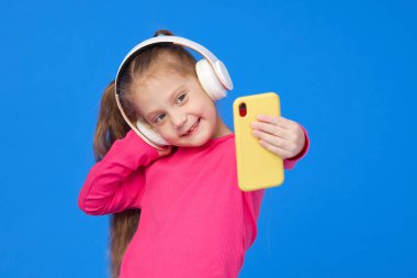 Parlak pembe kazaklı şirin bir kız cep telefonu kamerasıyla selfie çekiyor ve kablosuz kulaklıkla müzik dinliyor. Aletlerin üzerinde çocuk uygulamaları kavramı