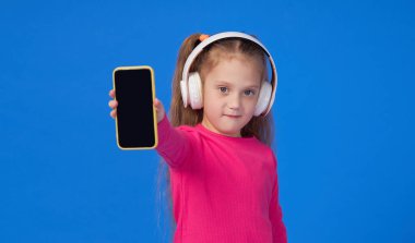 Parlak pembe kazaklı şirin bir kız elinde bir cep telefonu tutuyor ve kablosuz kulaklıkla müzik dinliyor. Aletlerin üzerinde çocuk uygulamaları kavramı