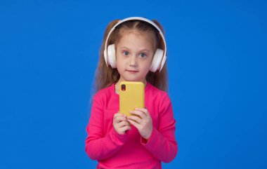Parlak pembe kazaklı şirin bir kız elinde bir cep telefonu tutuyor ve kablosuz kulaklıkla müzik dinliyor. Aletlerin üzerinde çocuk uygulamaları kavramı