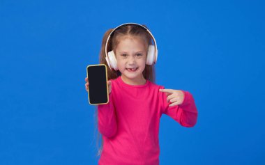 Parlak pembe kazaklı şirin bir kız elinde bir cep telefonu tutuyor ve kablosuz kulaklıkla müzik dinliyor. Aletlerin üzerinde çocuk uygulamaları kavramı