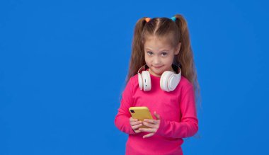 Parlak pembe kazaklı şirin bir kız elinde bir cep telefonu tutuyor ve kablosuz kulaklıkla müzik dinliyor. Aletlerin üzerinde çocuk uygulamaları kavramı
