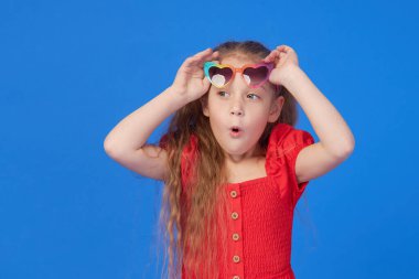 Kalp şeklinde güneş gözlüklü, şaşırmış, küçük kız çocuğunun portresi. Ağzı açık bir çocuk mavi arka planda eğleniyor. Kameraya bakıyorum. Komik surat.
